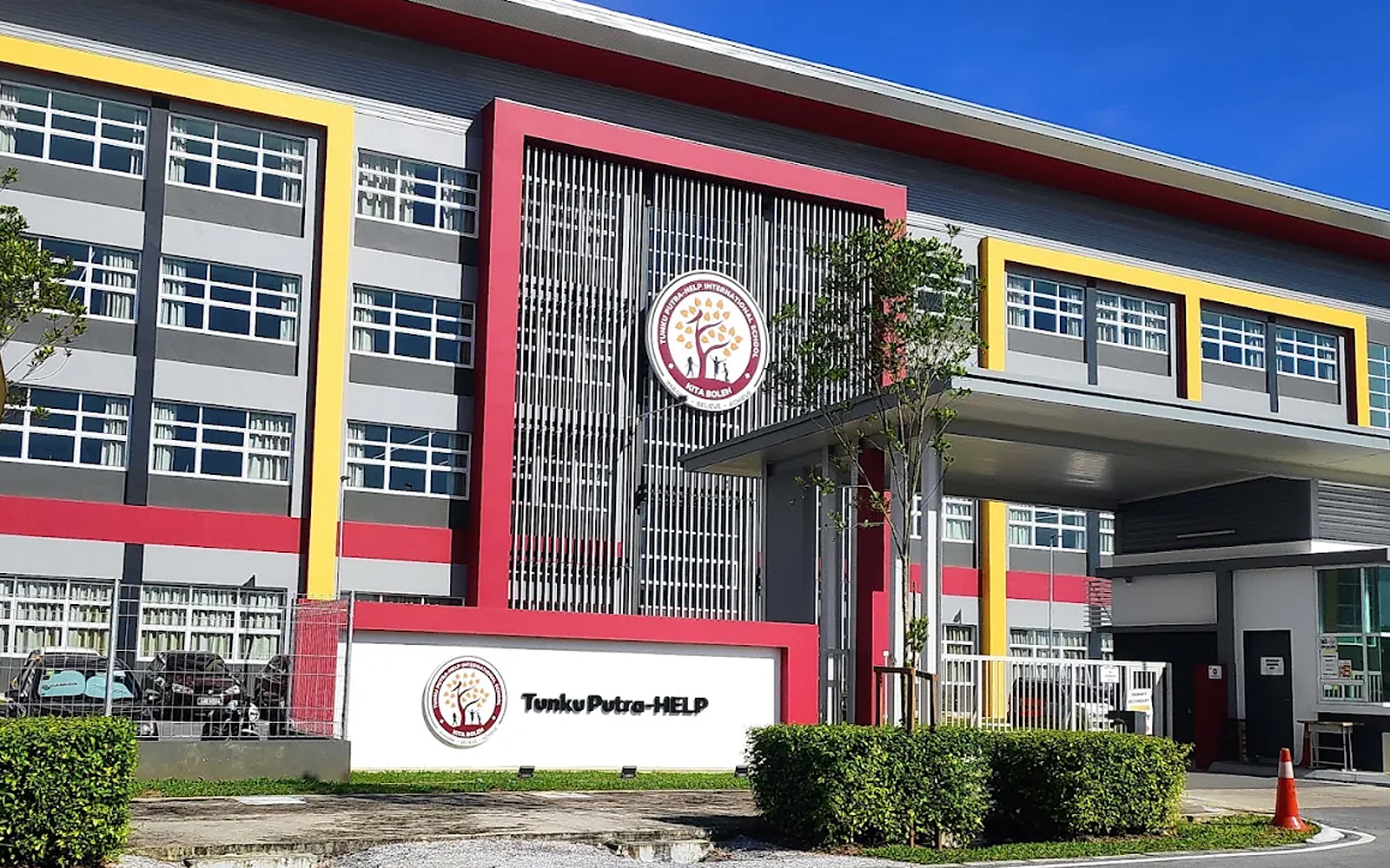 A grey, red and yellow multi-storey school building with a covered entrance and a Tunku Putra-HELP sign by the gate