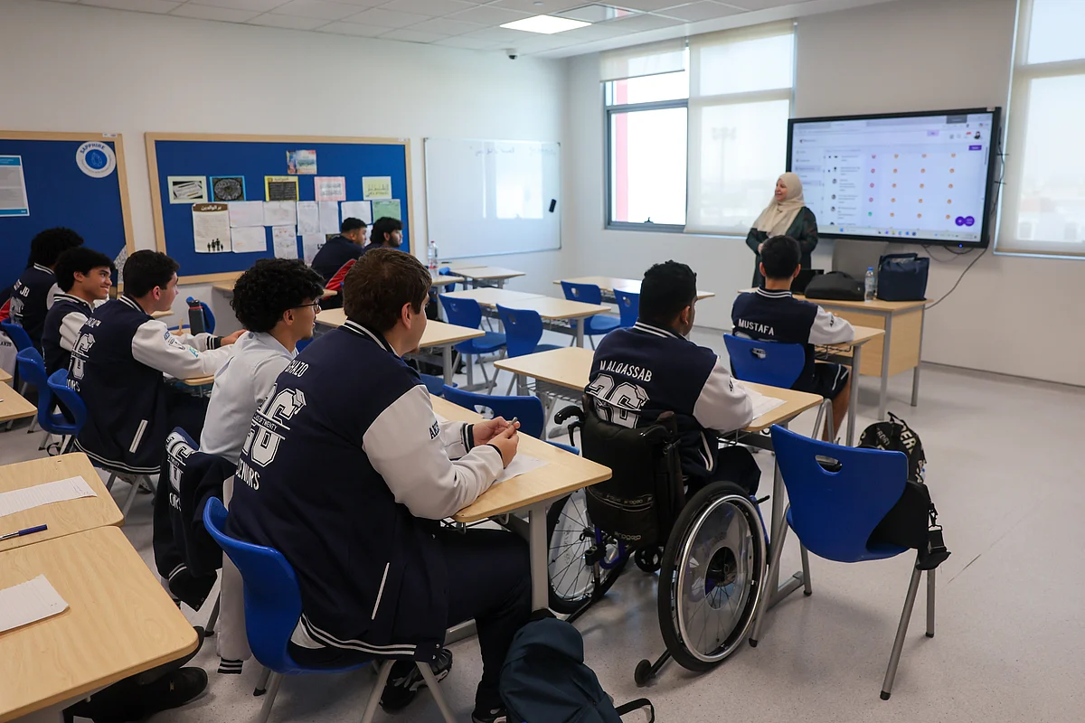 Teenage students in navy jackets sit at desks, one in a wheelchair, as a teacher stands by a large screen.