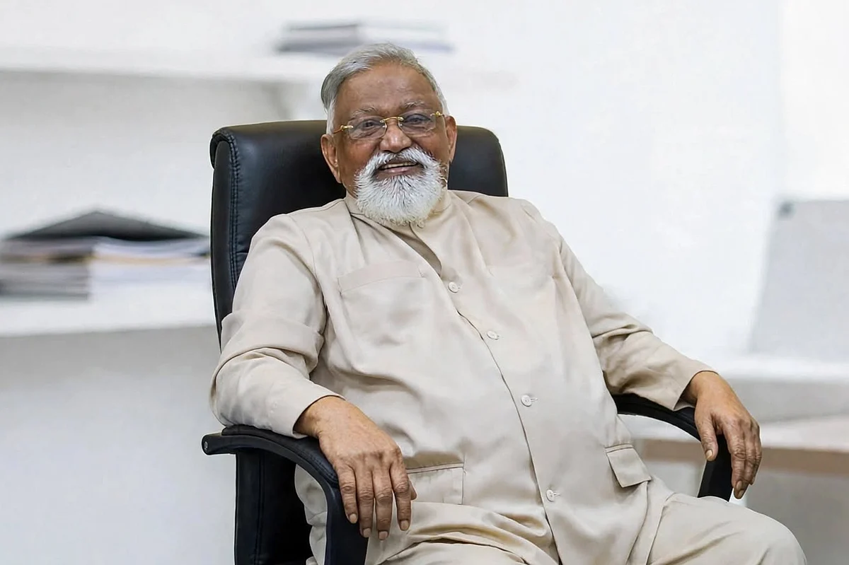 An older man with white hair, glasses and a white beard, in a beige tunic, smiling in a black office chair.