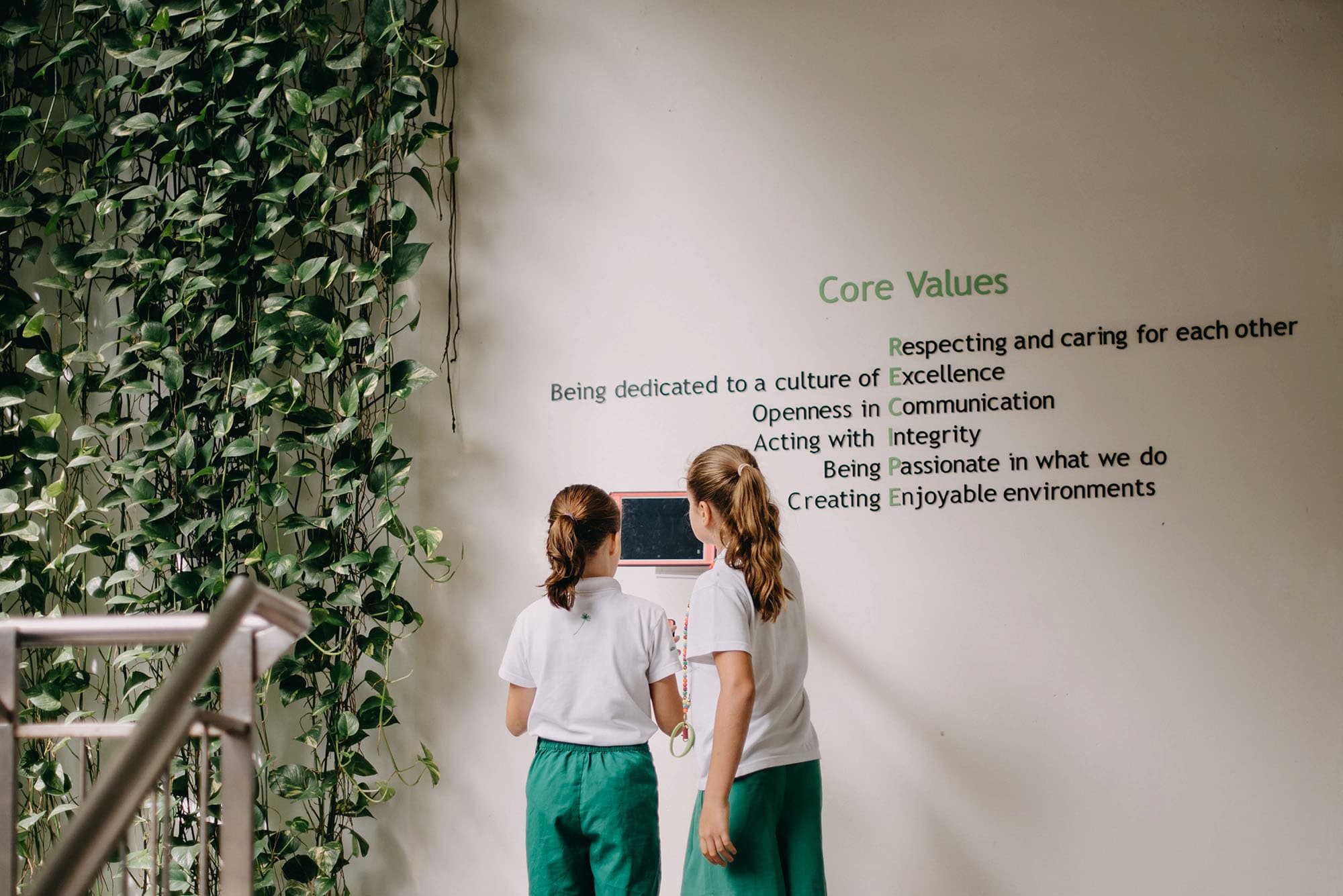 Two girls in white shirts and green trousers look at a wall-mounted tablet beside hanging plants and a values sign.