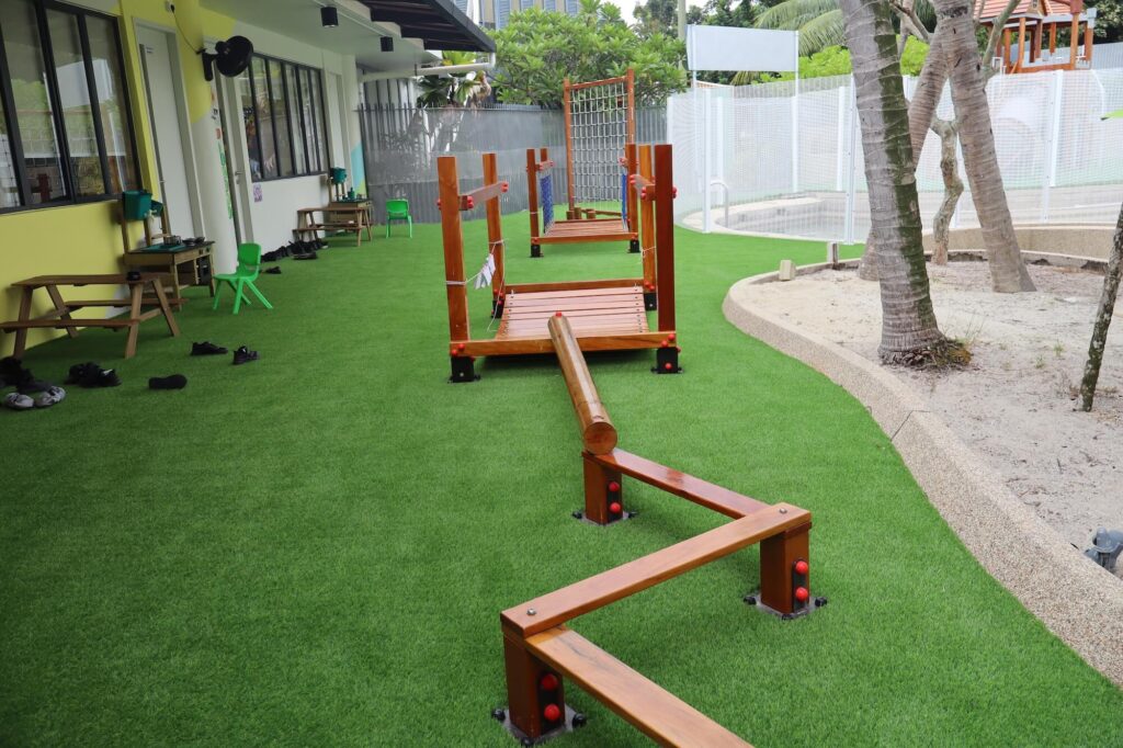 Wooden balance beams and a climbing frame on artificial grass beside a low school building with yellow walls.