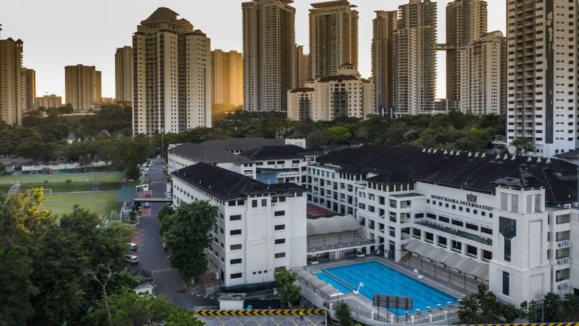 White school buildings with dark roofs, a swimming pool and playing fields, with tall apartment towers behind.