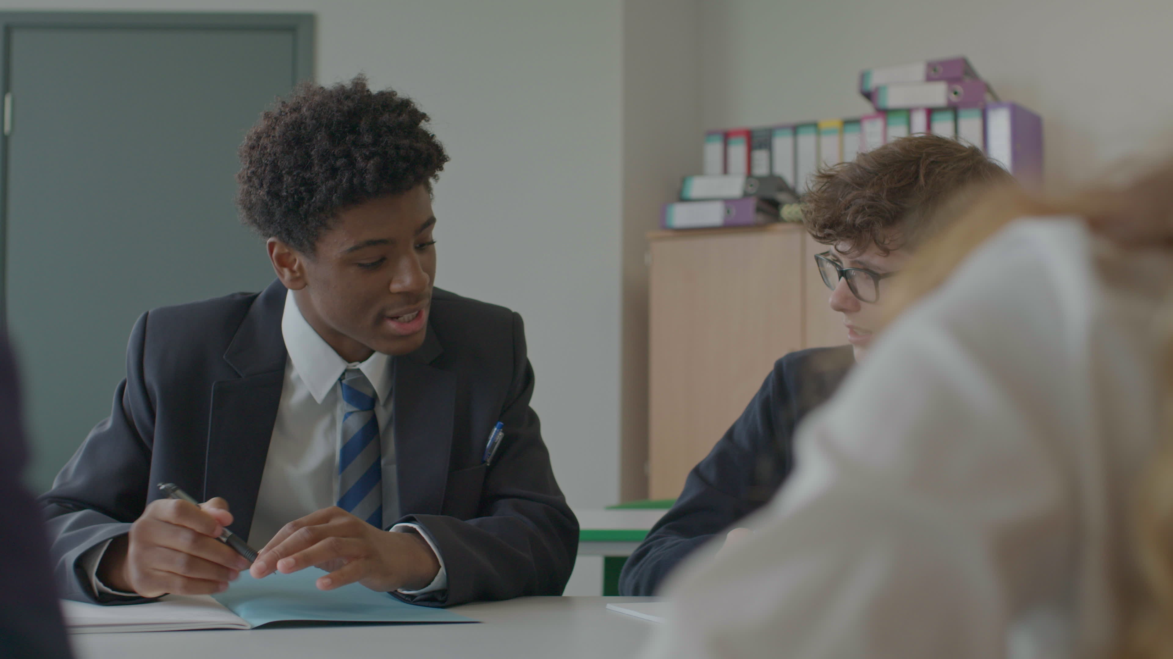 Two secondary pupils in blazers and ties talk over an open exercise book at a classroom desk, with folders on a shelf behind