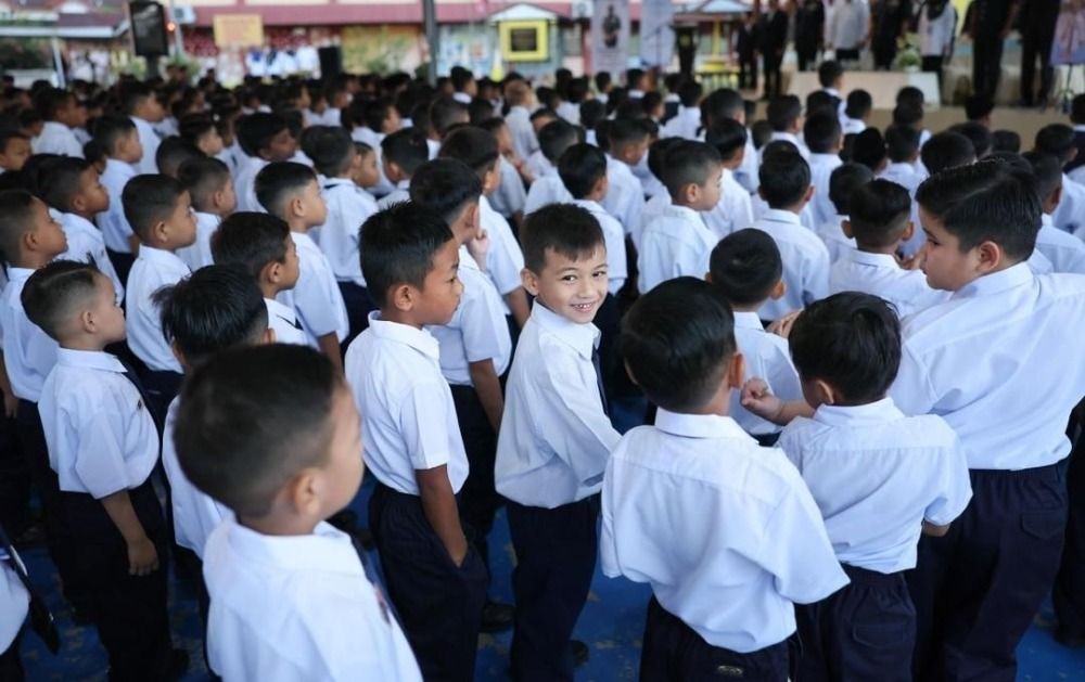 Rows of young boys in white shirts and navy trousers standing at a school assembly, one smiling back at the camera.