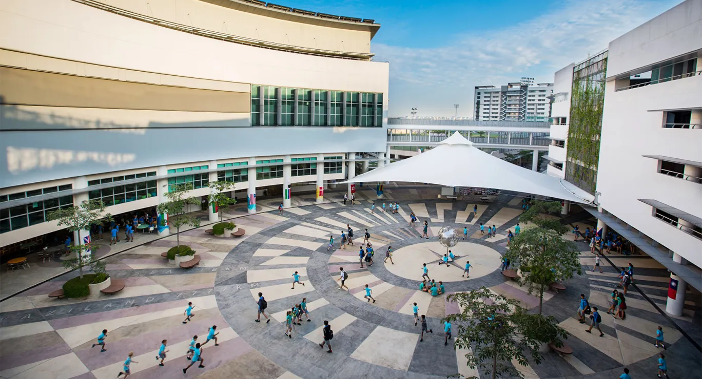 Pupils in blue shirts walk and run across a large paved courtyard between white school buildings under a shade sail.